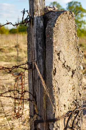 Weathered wooden and concrete fence post with rusted barbed wire. Dry grasses and a blurred field...