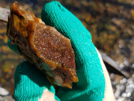 Orange druzy quartz specimen found in a sunny April stream, held by a green glove, reflecting water...