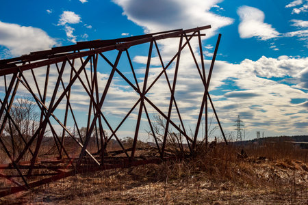 Rusted metal framework of a dismantled power line pylon lies on dry grassy terrain beneath a dramatic blue and white cloudy spring skyの写真素材