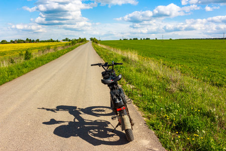 Fat tire electric bike parked on a narrow country road beside vibrant green fields under a bright blue sky with fluffy clouds: Russia - May 20, 2025のeditorial素材
