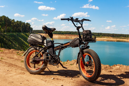 Rugged black fat tire electric bike parked on dirt overlooking bright blue quarry water and forest under summer sky: Russia - May 22, 2025のeditorial素材
