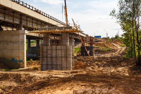 Under construction highway bridge with concrete pillars and scaffolding, heavy machinery and dirt mounds on the ground, bright sunny day: Russia - May 25, 2025のeditorial素材