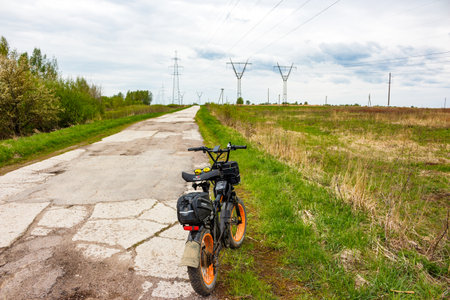 Rugged fat tire electric bike parked on a cracked country road amidst lush spring greenery and distant power line towers: Russia - May 5, 2025のeditorial素材