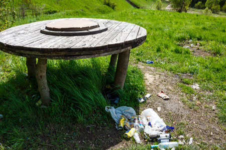 Rustic wooden table made from a spool sits in vibrant green grass surrounded by scattered trash: Russia - May 6, 2025のeditorial素材