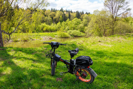 Black fat tire electric bike parked on vibrant green grass near a tranquil river and lush spring forest: Russia -May 7, 2025のeditorial素材