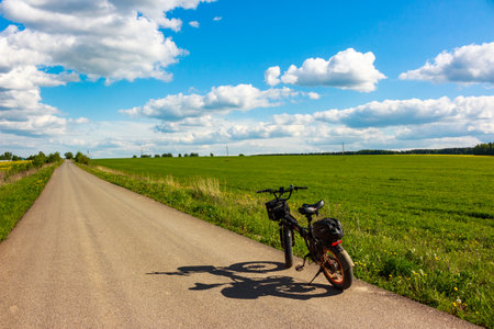 Black electric fat bike parked roadside next to a vibrant green field under a brilliant blue sky with puffy white clouds: Russia - May 20, 2025のeditorial素材