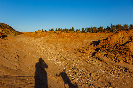 Photographer's shadow on a vast sand quarry floor. Orange earth and rock, clear blue sky, distant treesの写真素材