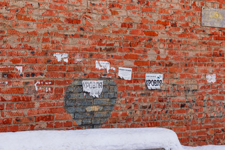 Weathered brick wall covered in peeling Russian roofing ads, contrasting with fresh winter snow....