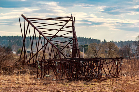 Deconstructed rusty metal electricity pylon lies fallen in a dry grassy field against a backdrop of distant trees and a cloudy spring skyの写真素材