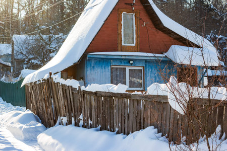 Rustic dacha house under a heavy blanket of fresh snow, sunlight illuminating the charming winter scene. A wooden fence in the foreground adds to the cozy, old-world vibeの写真素材