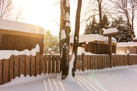 Golden hour illuminates a serene winter park. A rustic wooden fence draped in fresh snow defines the scene, with long shadows from bare trees stretching across the crisp, snowy groundの写真素材
