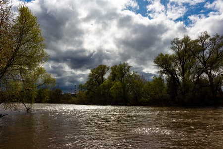 Glistening river flows beneath a dramatic, cloudy spring sky, framed by vibrant green trees....