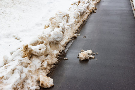 Grunge melting snow piled against a dark, wet asphalt path. Muddy slush and puddles signal the...