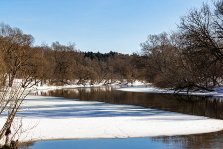 Melting river ice with open dark water reflecting bare branches under a bright blue spring sky