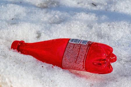 Vibrant red plastic bottle dumped in melting roadside snow. A poignant symbol of litter, waste, and...