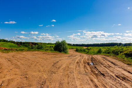 A bumpy dirt track winds through a vast open field under a bright blue summer sky with fluffy clouds, creating a scenic countryside travel route for off-road adventure