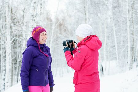 Two girls with a camera taking pictures in snow in winter in a park. Bright pink jacket. Photographer with the camera.の写真素材