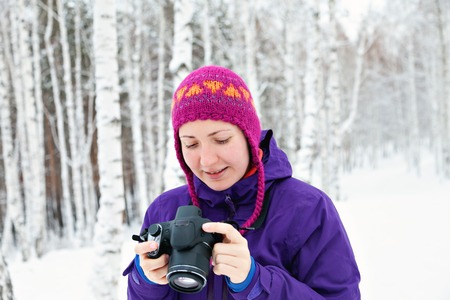 The girl with the camera on a background of snow in the winter woods. The bright colored hat and jacket. To watch photos. Photographer.の写真素材