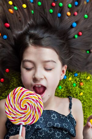 Little girl with dark hair lying on the floor among the sweets with his eyes closed. Top view of a...