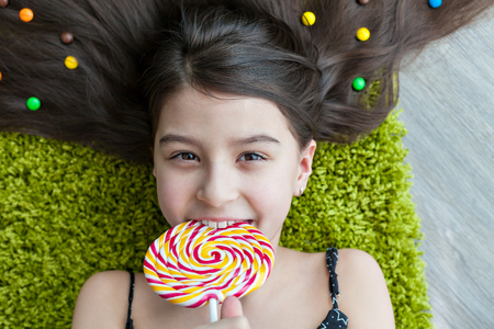 Little girl with dark hair lying on the floor among the sweets. Top view of a big yellow and pink lollipops in hands. Many candies in her hair. smile, joy and emotion on the face of the child.の写真素材