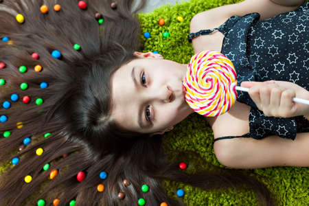 Little girl with dark hair lying on the floor among the sweets. Top view of a big yellow and pink lollipops in my mouth. Many candies in her hair. fun and excitement on the face of the child.の写真素材