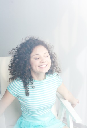 Portrait of a young girl sits in a white chair. Turquoise dress and wavy hair.の写真素材