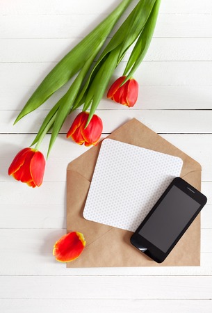 Spring bouquet of red tulips and black mobile phone on a white wooden background with copy space. View from the top with space for signature.の写真素材