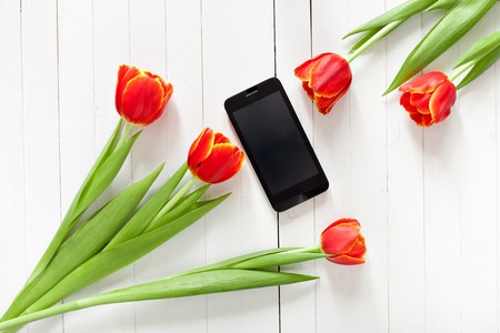 Spring bouquet of red tulips and black mobile phone on a white wooden background with copy space. View from the top with space for signature.の写真素材