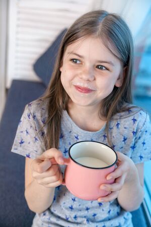 Closeup portrait of a cute little girl with a white milk mustache, pleased charming child with a smile holds a glass, drinks milk before bedtimeの写真素材