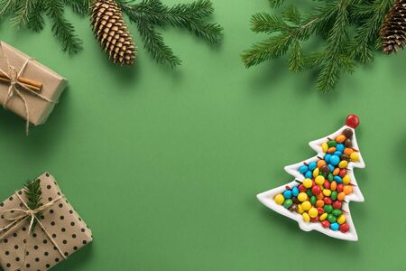 Christmas composition. Gifts and branches of spruce. A plate in the form of a Christmas tree filled with colorful candies on a green background. Flat lay, top view, copy spaceの写真素材