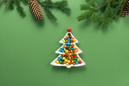 Christmas composition. A plate in the form of a Christmas tree filled with colorful candies and branches of spruce on a green background. Flat lay, top view, copy space.の写真素材