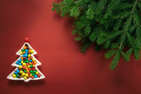 Christmas composition. A plate in the shape of a Christmas tree filled with colorful candies. Branches of spruce on a red background. Flat lay, top view, copy space.の写真素材