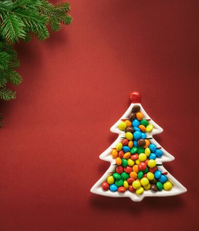Christmas composition. A plate in the shape of a Christmas tree filled with colorful candies. Branches of spruce on a red background. Flat lay, top view, copy space.の写真素材