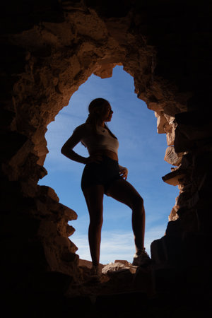 A silhouette of a young athletic woman framed by the natural rock edges of a cave looking confidently toward the horizon under a clear blue sky. This empowering image captures the essence of adventure strength and exploration perfect for themes related to outdoor sports travel nature and female empowermentの写真素材