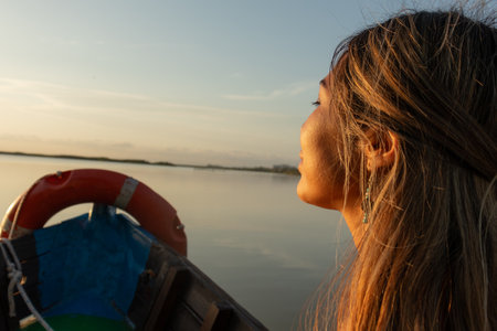 Young Latina woman with dark hair gazing at the sunset from a boat on a serene lake. Natural light illuminates her face creating a warm and tranquil atmosphere while the girl with an expression of awe and serenity seems deeply in love with the landscape. The scene conveys a sense of peace freshness and romance evoking the spirit of a relaxing summer afternoon.の写真素材