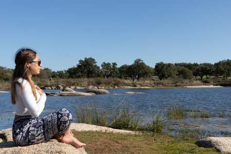 A young woman sits peacefully on a rock by a tranquil lake with her hands placed over her heart in a meditative pose. Her calm expression and serene surroundings reflect a deep connection with nature and inner peace. This image symbolizes the soul's journey mindfulness and self-reflection capturing the essence of calm balance and introspection.の写真素材