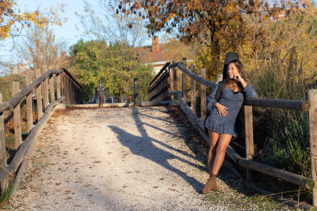 Woman looking placidly to her right with a blue dress showing her legs brown boots and a guitar in its case on her back leaning with one arm on the railing of a wooden and metal bridge and the other arm resting on her abdomen her shadow projected in autumn with lots of yellow and red green vegetation some houses in the background and a road of small stones.の写真素材