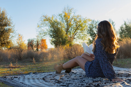 beautiful woman with brown and golden hair in a blue dress with brown bonnets reading a book sitting on a mandala in nature reading with one hand while holding the book with the other surrounded by nature and autumn colored trees.の写真素材