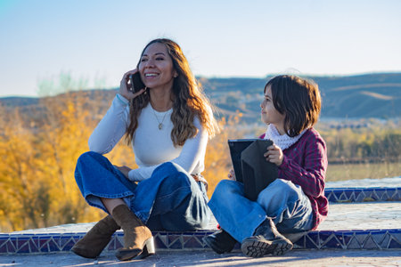 A beautiful mother smiles as she sits and talks on the phone while her young daughter looks up at her with admiration and love. The little girl is holding a tablet but her attention is focused on her mother. It reflects emotional well-being family connection and the joy of spending quality time together outdoors.の写真素材