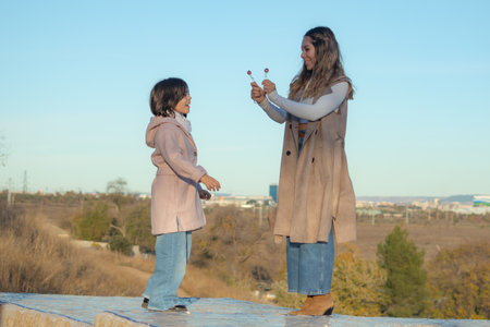 A young mother wearing a long brown coat is happily revealing two lollipops she had hidden to her young daughter in a beautiful autumn park. Surrounded by trees with vibrant fall foliage, the little girl looks at the lollipops with wide eyes, full of surprise and excitement.の写真素材
