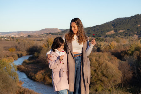 A young, beautiful mother and her daughter are embracing while enjoying lollipops together by a tranquil river surrounded by the warm autumn colors. The mother looks at her daughter with love and tenderness while the little girl is savoring her lollipop clearly happy and content.の写真素材