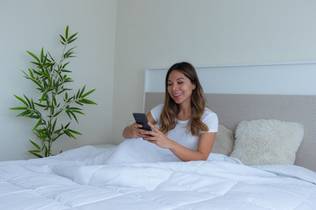 pretty latin woman in a white bed with soft cushions in a quiet room looking and typing on the mobile phone just up relaxed and enjoying a moment of relaxation with a green plant in the background, and a beige headboard in a quiet and peaceful environment.の写真素材