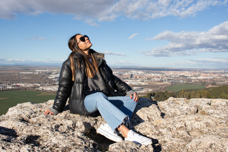 A young woman with golden hair, jeans and white sneakers. She is sitting on a rock on a hill, enjoying the sun with her sunglasses and with views of the city in the background.の写真素材