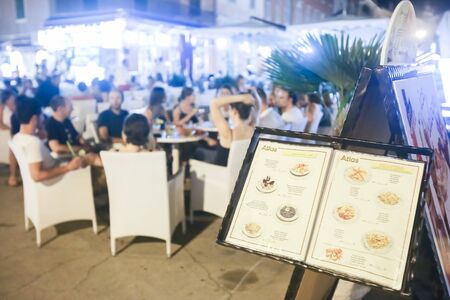 ROVINJ CROATIA  AUGUST 6 : A restaurant menu displayed on a stand with people sitting in a restaurant on the promenade at night on August 6th 2013 in Rovinj Croatia. Rovinj is a tourist destination on Adriatic coast of Croatia.のeditorial素材