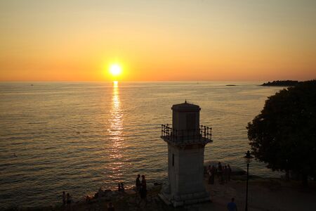 A group of tourists standing on the shore by the lighthouse and watching the sunset on in Rovinj, Croatia.の写真素材