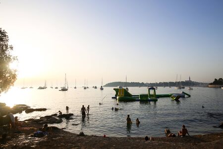 ROVINJ, CROATIA - AUGUST 3 : Tourists sunbathing and swimming in the Adriatic sea with a group of sailboats anchored at sunset on August 3rd, 2013 in Rovinj, Croatia.のeditorial素材