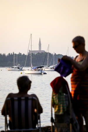 ROVINJ, CROATIA - AUGUST 3 : A man sitting on a chair and a woman standing next to him on the shore with boats anchored in the Adriatic sea in front of the Saint Eufemia church on August 3rd, 2013 in Rovinj, Croatia.のeditorial素材