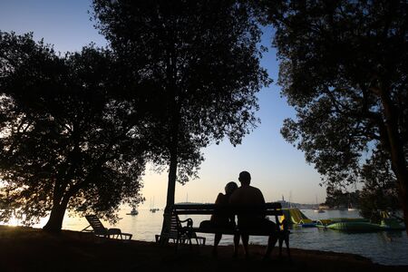 ROVINJ, CROATIA - AUGUST 3 : A rear view of a couple sitting on a wooden bench on the shore and watching the sunset on Adriatic sea on August 3rd, 2013 in Rovinj, Croatia.のeditorial素材