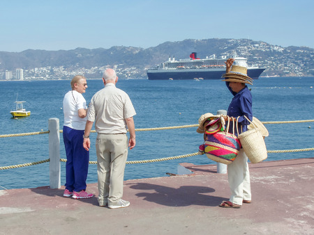 ACAPULCO, MEXICO - MARCH 11, 2006 : A Mexican selling hats to tourists with Queen Mary 2 cruise ship in the background in Acapulco, Mexico.のeditorial素材