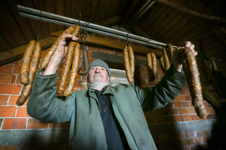 A front view of an old man posing with dry sausages hanging in the attick.の写真素材